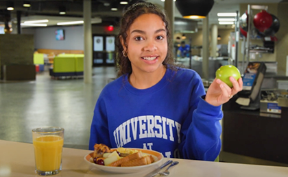 Female student wearing a University at Buffalo sweatshirt holding an apple and eating brunch and orange juice at a table at Crossroads Culinary Center (C3).