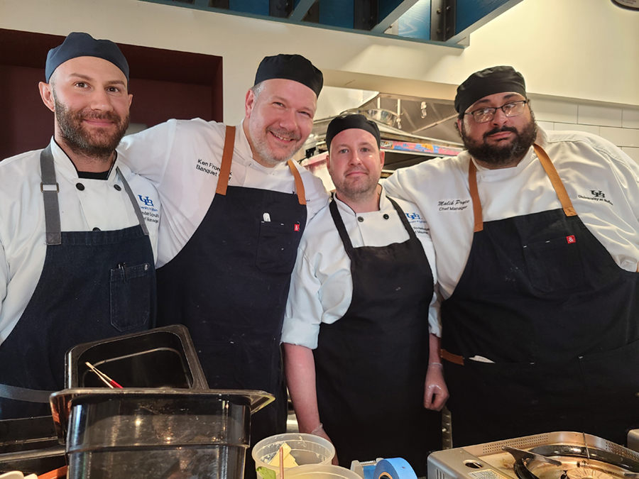 The four chefs posing and smiling at the camera while making the dishes