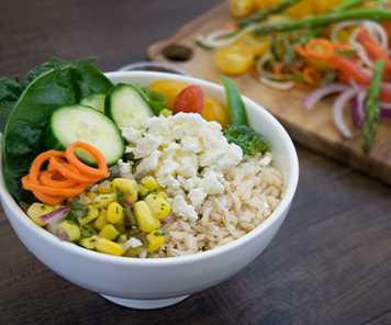 Bowl consisting of rice, feta cheese, roasted corn, onions, cucumber, carrots, tomatoes, broccoli and romaine lettuce.