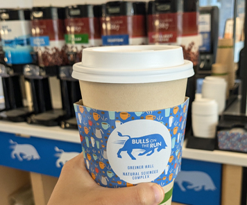 A large hot cup of coffee placed on a table with Bulls on the Run Greiner in the background