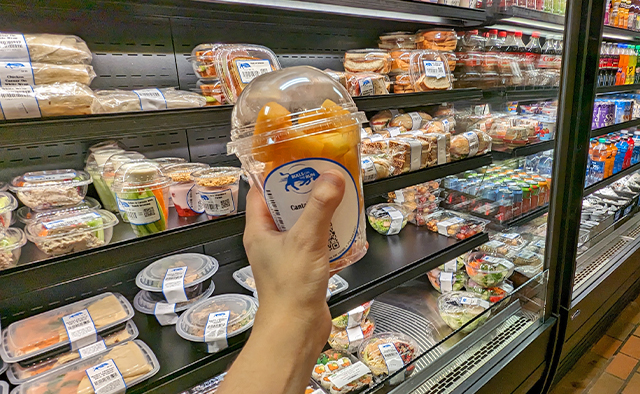 Hand holding up a Grab and Go fruit container in front of the shelves full of Grab and Go offerings