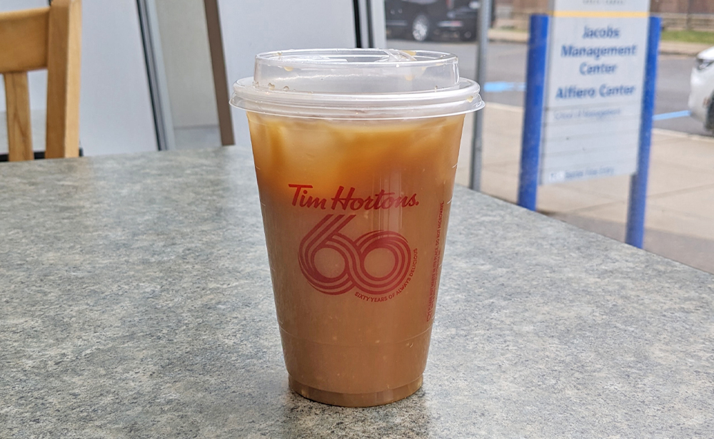 an iced Tim Hortons coffee on a table, with a sign outside the window that reads Jacobs Management Center Alfiero Center