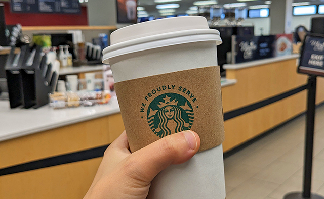 A hot coffee with the Proudly Serve Starbucks logo in the foreground, with Whispers Café in Capen Hall in the background