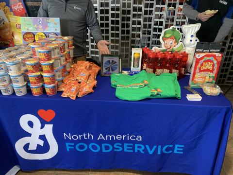 Table with a General Mills table cloth, and General Mills cereal cups on the top, alongside Lucky Charms T shirts and water bottles.