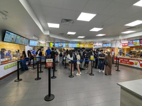 A view of Union Marketplace from the entrance, showing all stations from left to right: Union Station, Halal Shack, Piza Pizza, and Fowl Play. Customers are in line at every station.