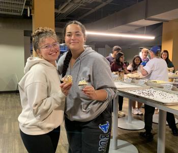 Two students smiling and showing their decorated cupcakes, with people decorating baked goods in the background