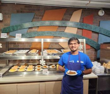 Associate posing with a plated pumpkin pie slice, in front of the other desserts at Governors Dining Center