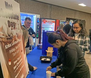 Student scooping marshmallows onto their hot chocolate at the table, with a line forming behind them.