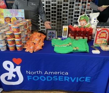 Table with a General Mills table cloth, and General Mills cereal cups on the top, alongside Lucky Charms T shirts and water bottles.