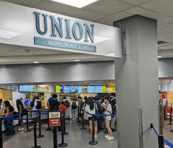 A view of Union Marketplace from the entrance, showing the logo sign above the entrance, and the stations and queue of customers are past the sign.