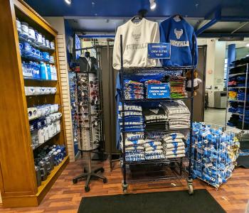 Shelving in the Campus Tees store full of mugs, tumblers, t-shirts and crew sweatshirts. There's a sunglass display as well, and a bin of Victor E. Bull plushies by the door.