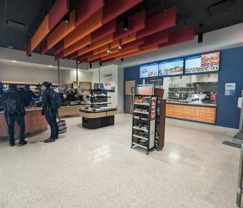 Customers checking out at Corner Café. You can see some grab & go items, a snack shelf, the full digital menuboards above the associates preparing the food.