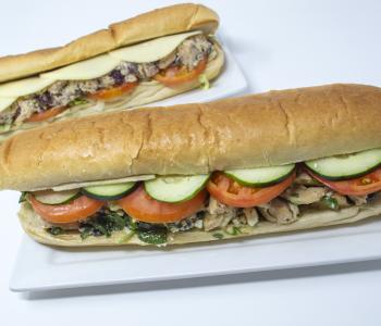 Two chicken subs from Union Station. The back one is topped with provolone cheese, tomatoes, and lettuce. The front one is topped with cucumbers, tomatoes and spinach.